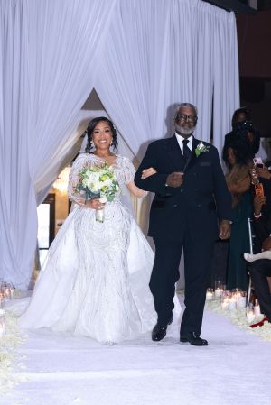 A bride in a white, beaded wedding gown walking down the aisle with her father, who is wearing a black tuxedo, at their wedding ceremony.