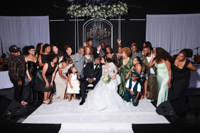 A large wedding party gathered around the bride and groom on a stage decorated with flowers and a chandelier.