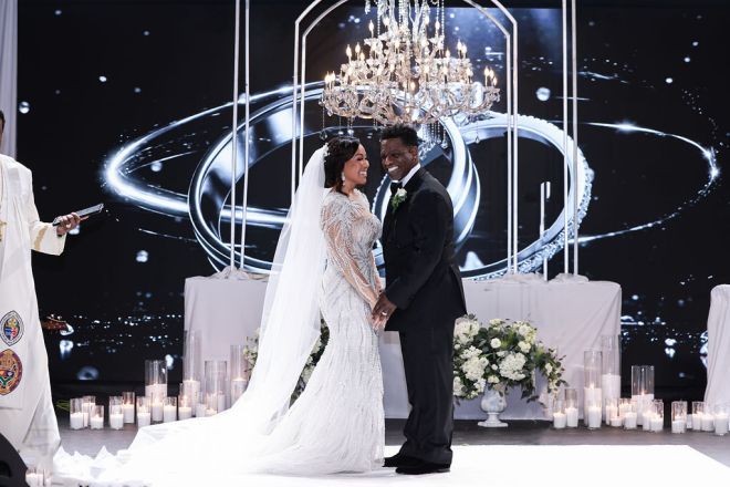 A bride and groom standing together in a lavish wedding setting, with a large crystal chandelier and glowing candles surrounding them.
