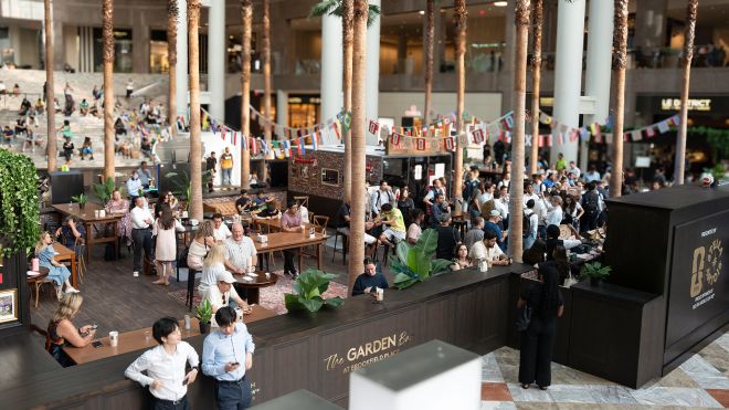 Crowded indoor dining area with palm trees, flags, and people seated at tables in a lively atmosphere.