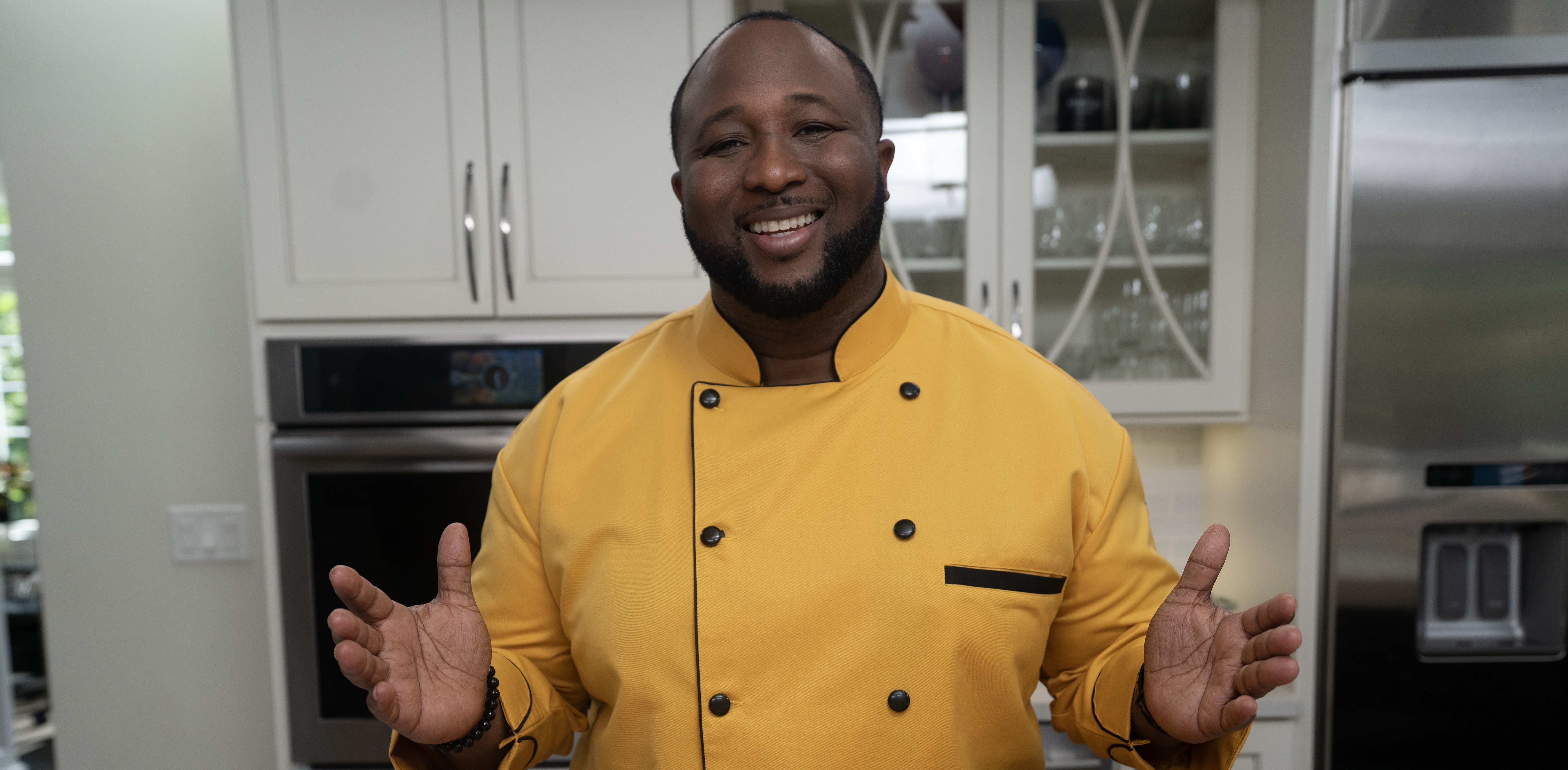 A smiling man in a yellow chef's jacket standing in a kitchen, with various ingredients and cooking tools on a counter in front of him.