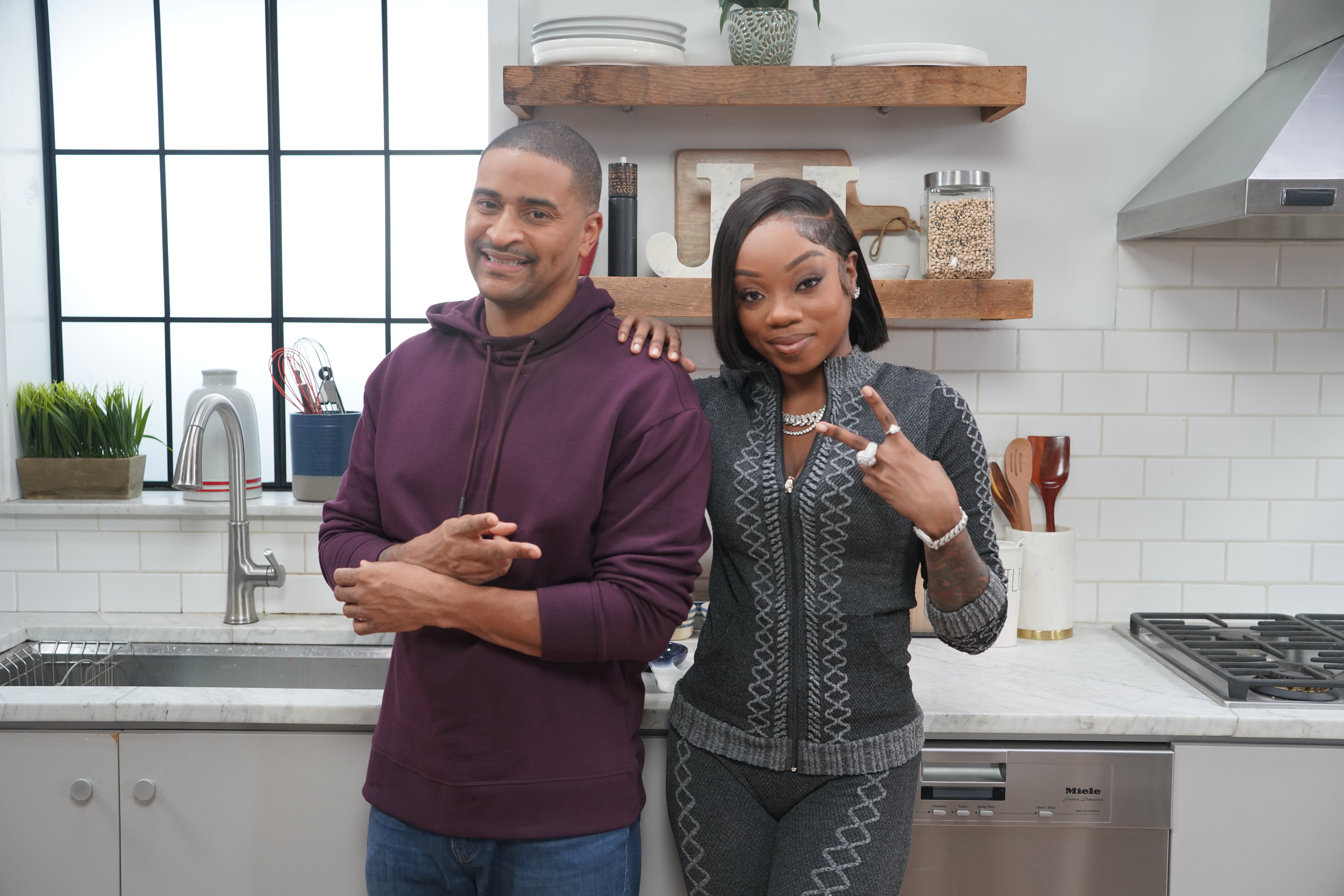 A smiling man and woman standing together in a kitchen, with shelves and appliances visible behind them.