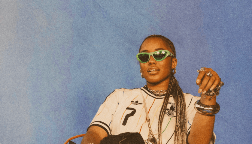 A person wearing sunglasses, jewelry, and a jersey with the number 7, posing against a blue background.