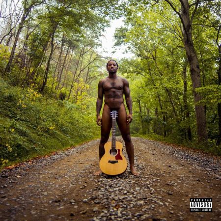 A shirtless man holding an acoustic guitar stands on a dirt path in a lush, green forest.