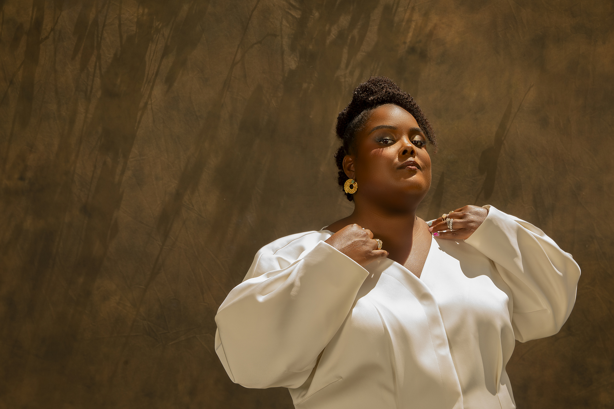 A woman in a white blouse stands with her arms crossed, looking thoughtfully upward against a warm-toned background.