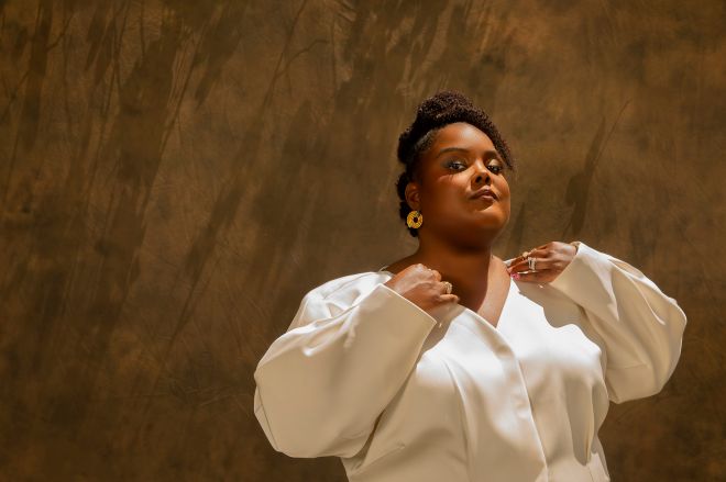 A woman in a white blouse stands with her arms crossed, looking thoughtfully upward against a warm-toned background.