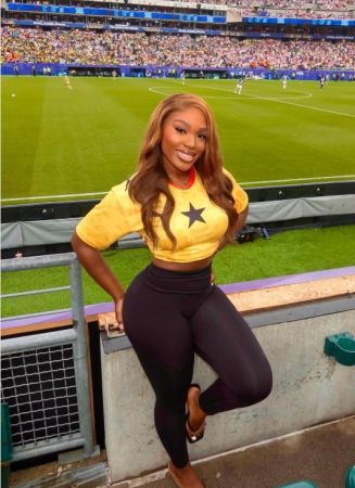 A woman wearing a yellow jersey with a star logo stands in a sports stadium, smiling at the camera.