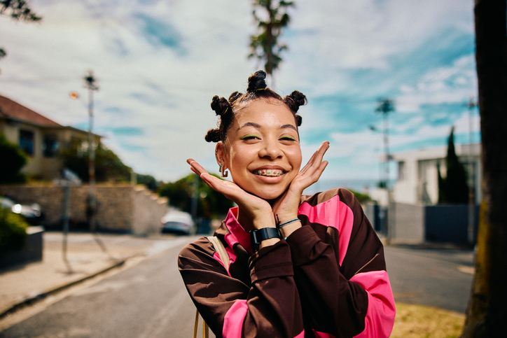 Eccentric teen girl with bantu knots and braces smiling at camera with hands on cheeks in street