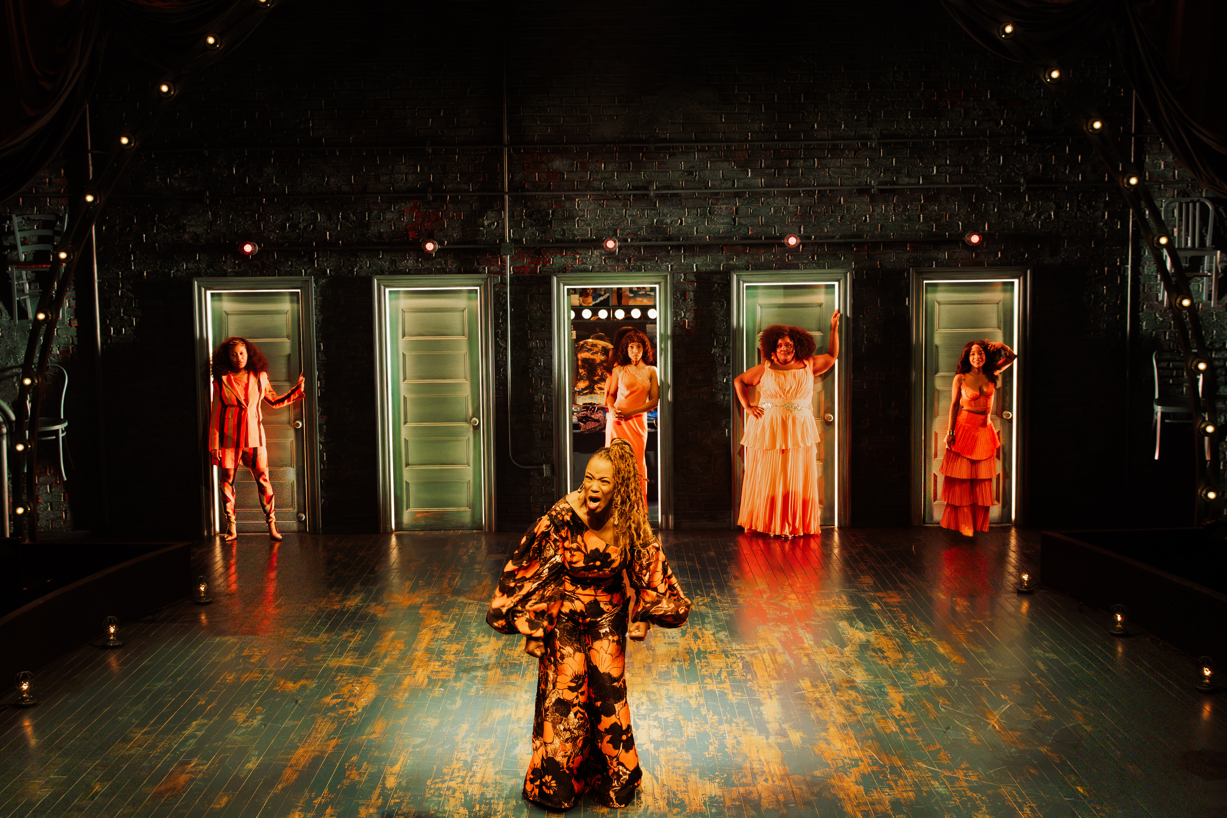 A performer in a colorful, patterned dress dances on a stage with several other performers in the background, all illuminated by warm lighting.