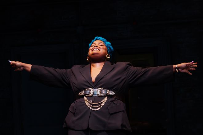 A person with blue hair standing with arms outstretched on a dark stage.