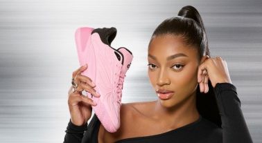 A woman in a black crop top and skirt holding a pink sneaker, posing in front of a gray background.