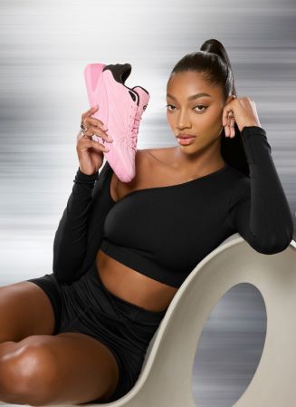 A woman in a black crop top and skirt holding a pink sneaker, posing in front of a gray background.