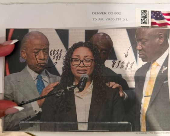 Three African American individuals, two men and one woman, standing at a podium and speaking into microphones. The woman is wearing glasses and a black jacket. The background includes an American flag.