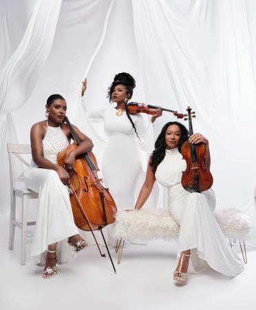 Three women in white dresses holding violins and cellos performing on a white stage with draped curtains.