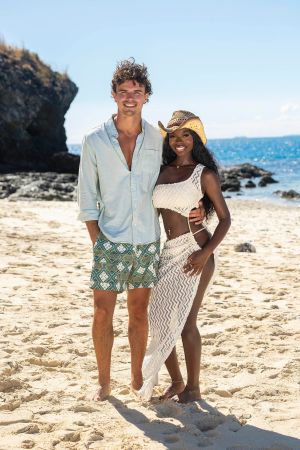 A smiling couple on a beach, the man wearing a light blue shirt and the woman wearing a white and tan patterned dress and cowboy hat.