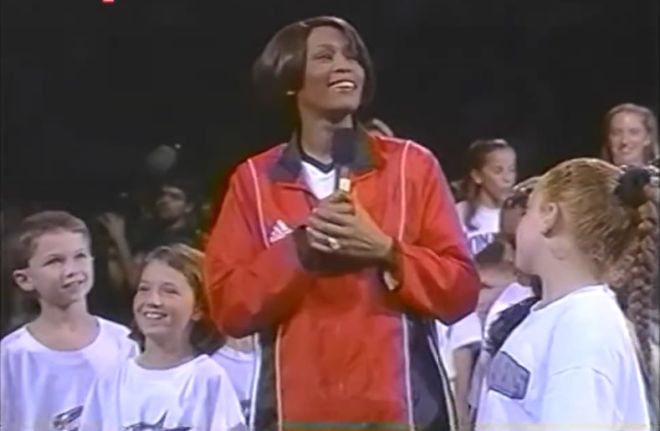 A person wearing a red Adidas jacket standing on a stage and smiling while surrounded by a crowd of people.