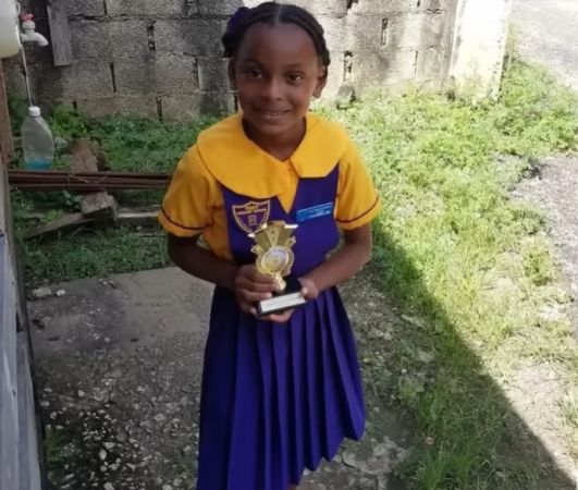 A young girl in a yellow and purple school uniform holding a trophy and smiling in front of a stone wall.