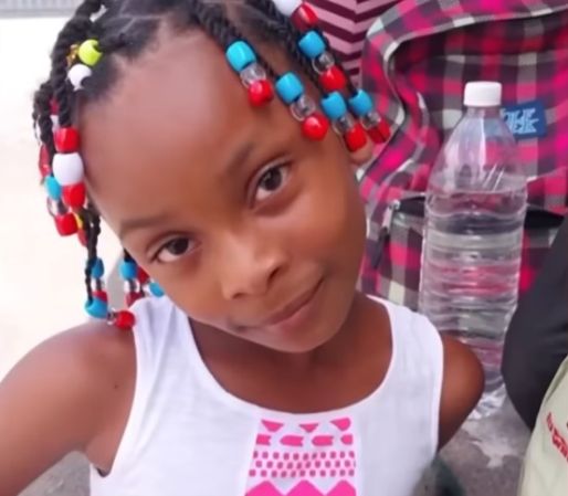A young girl with braided hair and colorful beads, wearing a white tank top with a pink graphic design.
