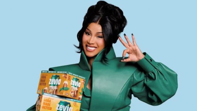 A smiling woman in a green jacket holding boxes of Zevia soda, making a peace sign gesture.
