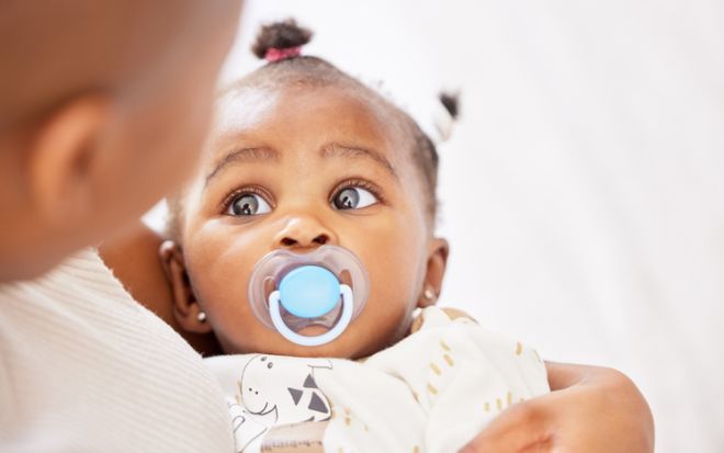 Shot of an adorable baby girl sucking a dummy while being held by her mother at home