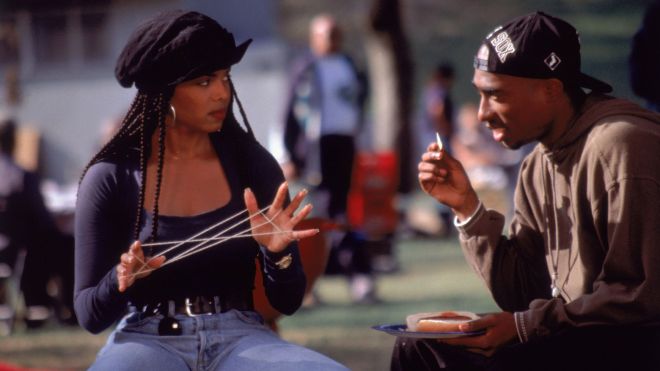 Two people, a woman with braided hair and a man wearing a cap, engaged in conversation outdoors.