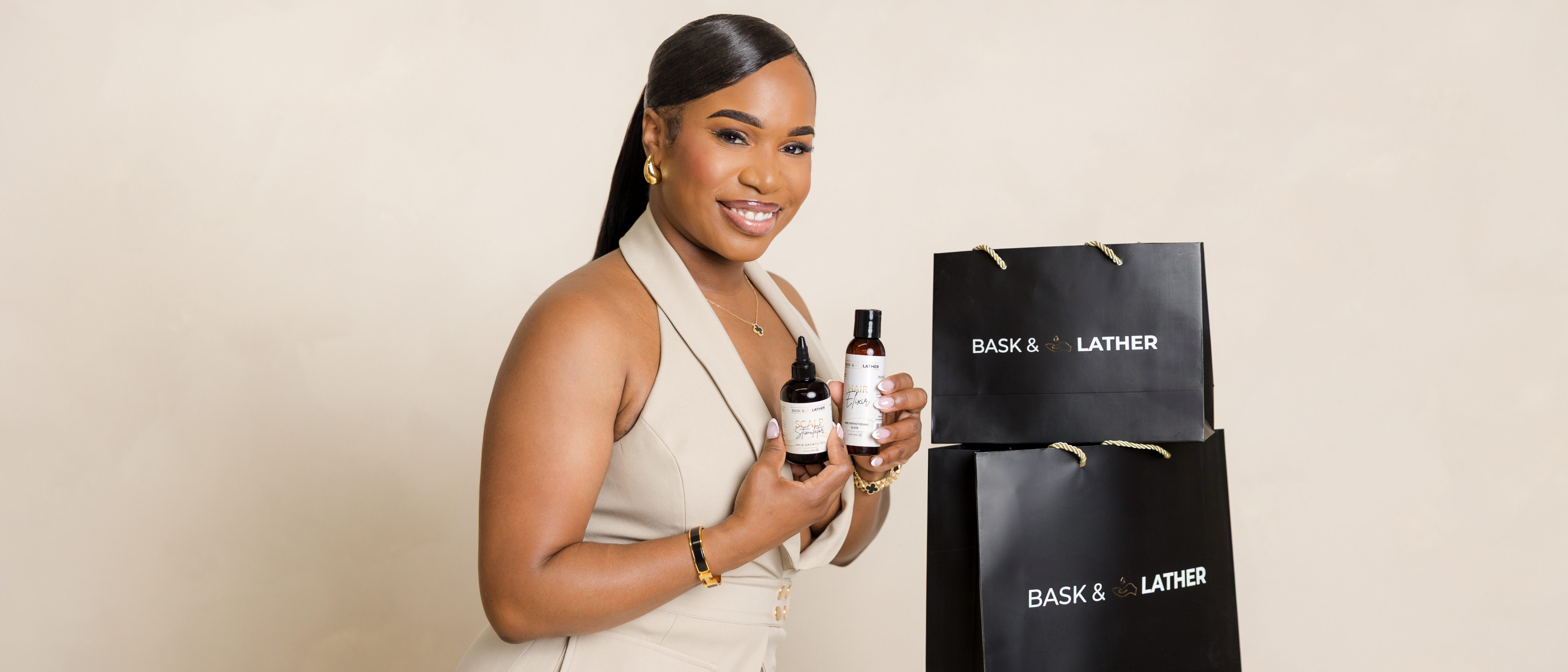 A smiling woman in a beige suit holding two bottles of Bask & Lather skincare products in front of a display of the same products.