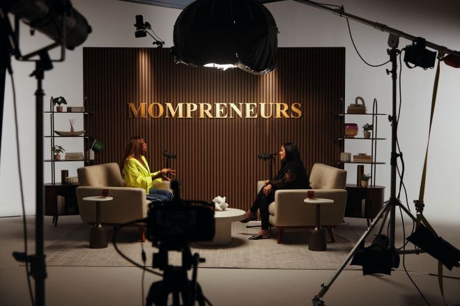 Two women sitting in chairs in a studio setting, with "MOMPRENEURS" displayed prominently on the background.