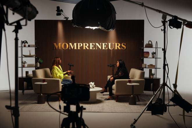 Two women sitting in chairs in a studio setting, with "MOMPRENEURS" displayed prominently on the background.