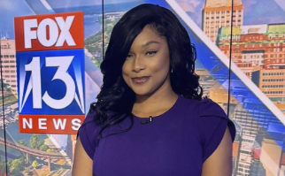 A woman in a purple dress standing in front of a Fox 13 News backdrop with a cityscape visible through the studio windows.