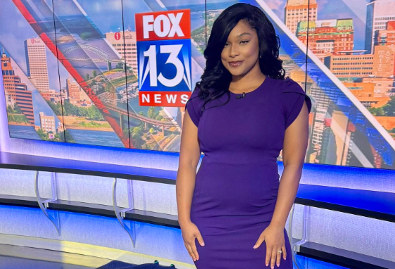 A woman in a purple dress standing in front of a Fox 13 News backdrop with a cityscape visible through the studio windows.