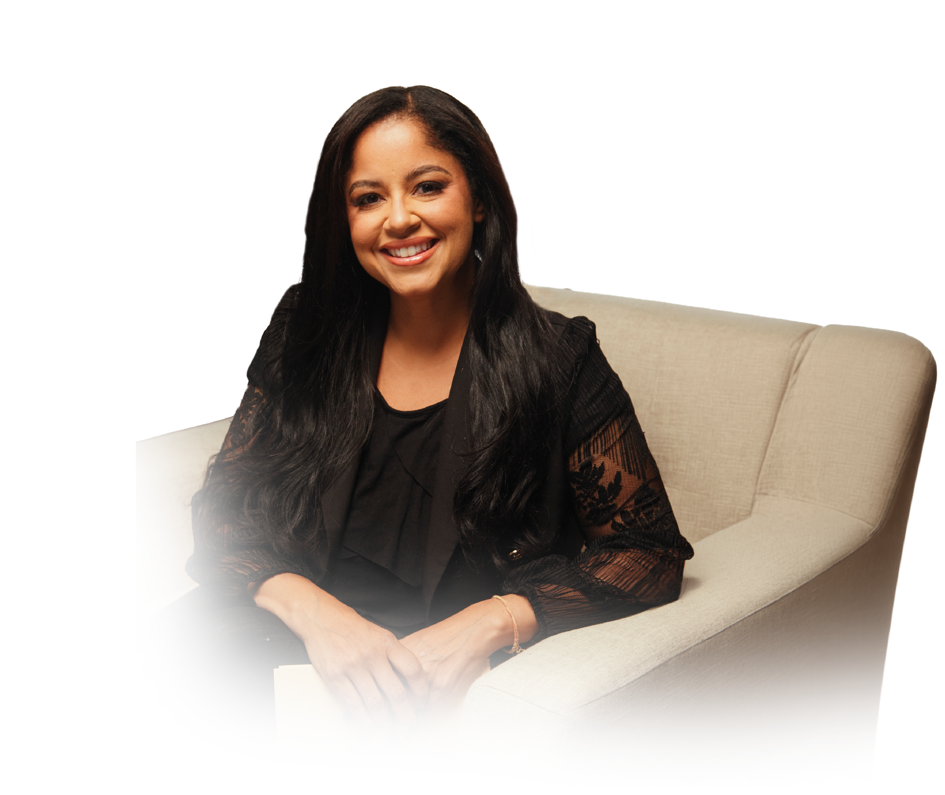 A smiling woman with long dark hair wearing a black lace top and sitting on a beige couch.