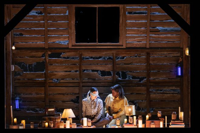 Two people sitting on the floor of a rustic wooden cabin, surrounded by lit candles.