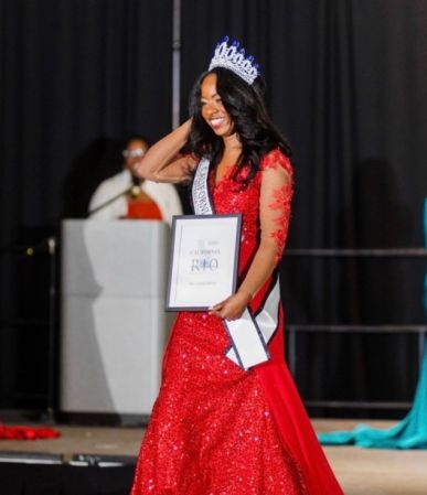 A woman wearing a red sequin dress and a crown stands on a stage holding a framed award.