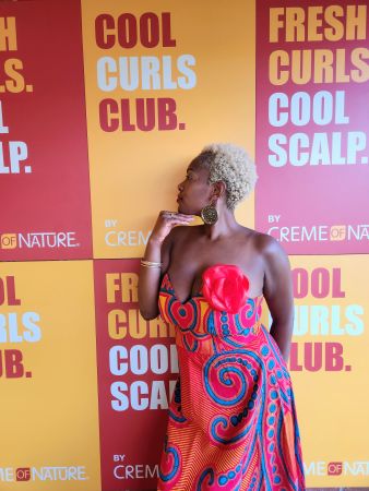 A woman with curly gray hair wearing a colorful patterned dress stands in front of signs for "Cool Curls Club" and "Fresh Curls Cool Scalp" by Creme of Nature.