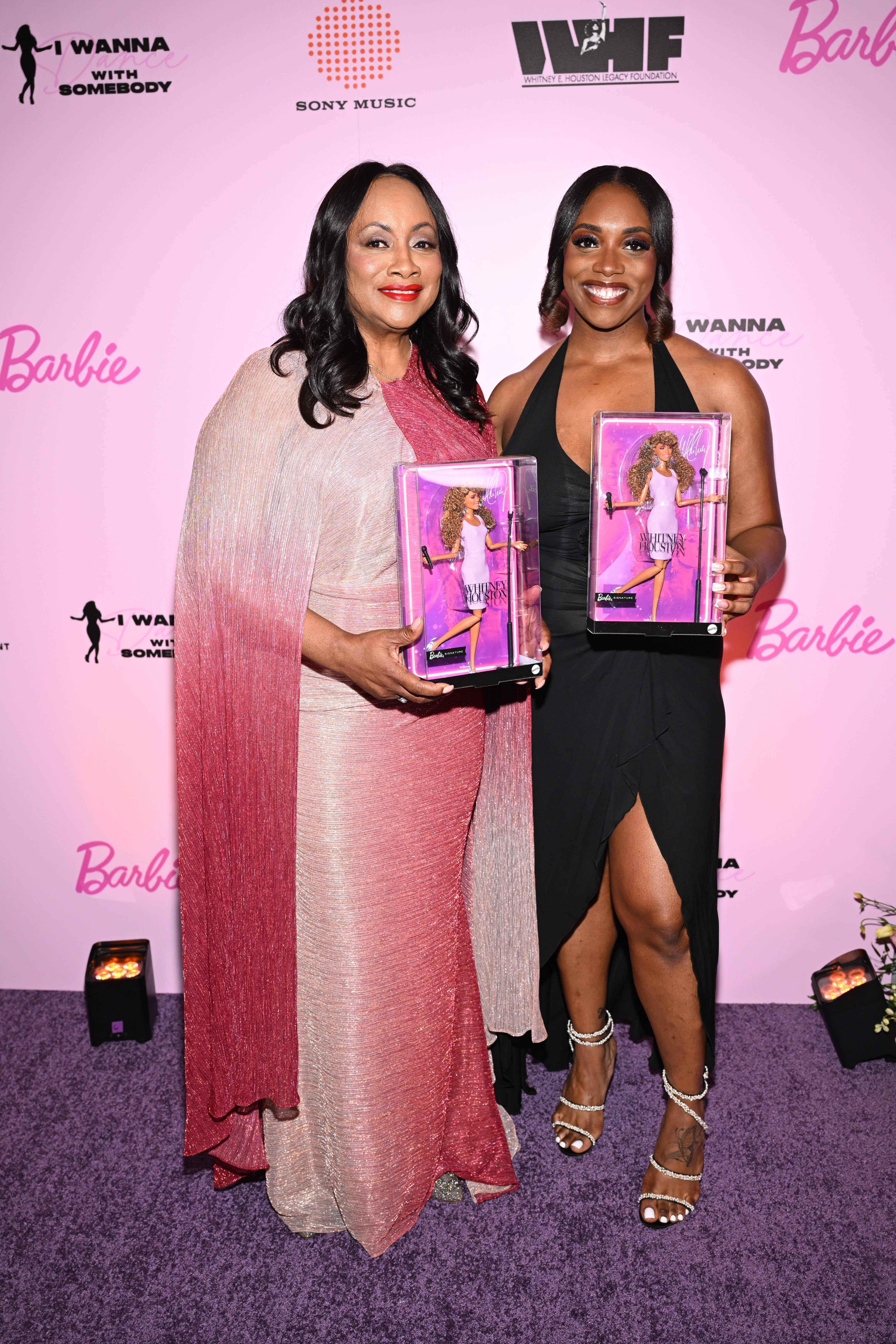 Two women holding Barbie dolls posing in front of a pink backdrop with Barbie and "I Wanna With Somebody" branding.