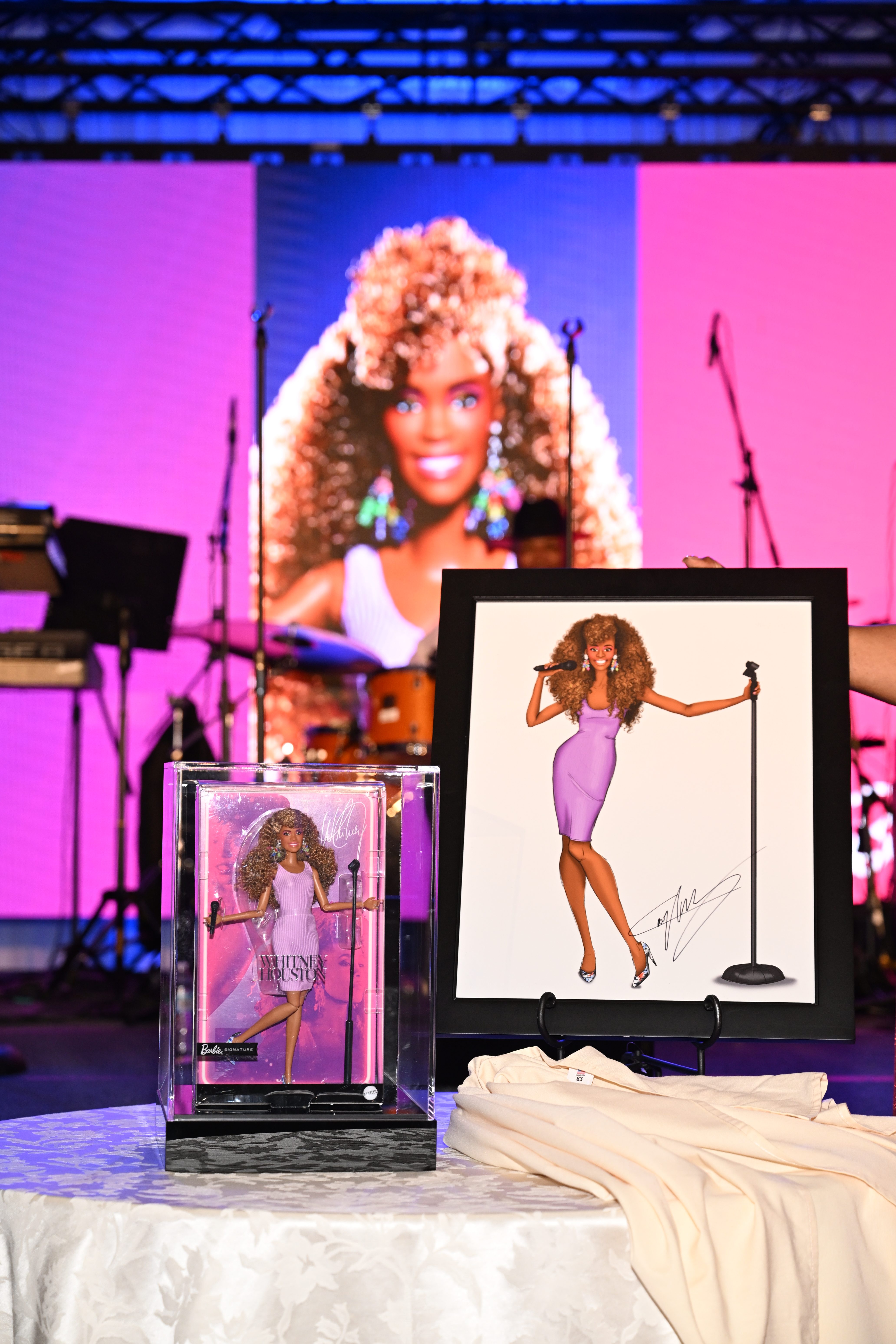 A stage with a large screen displaying a close-up image of a woman with curly red hair wearing a pink dress and singing into a microphone. In the foreground are a framed illustration of the same woman and a Barbie doll in a similar outfit.