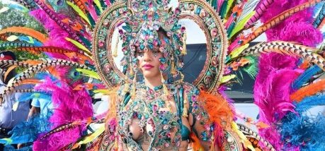 A person wearing an elaborate, colorful costume with feathers, beads, and jewels, standing in front of a crowd at what appears to be a carnival or festival.