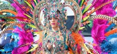 A person wearing an elaborate, colorful costume with feathers, beads, and jewels, standing in front of a crowd at what appears to be a carnival or festival.