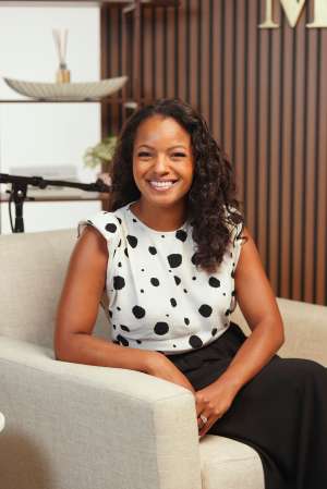 A smiling woman with curly hair wearing a polka dot top and sitting on a couch in a room with wooden paneling.