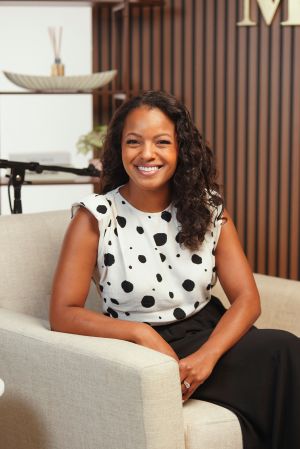 A smiling woman with curly hair wearing a polka dot top and sitting on a couch in a room with wooden paneling.