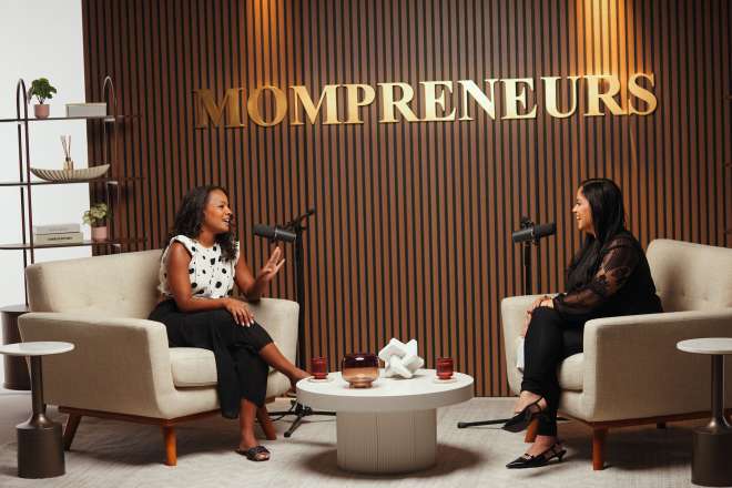 Two women seated in chairs, engaged in conversation, with the text "MOMPRENEURS" displayed on the wall behind them.