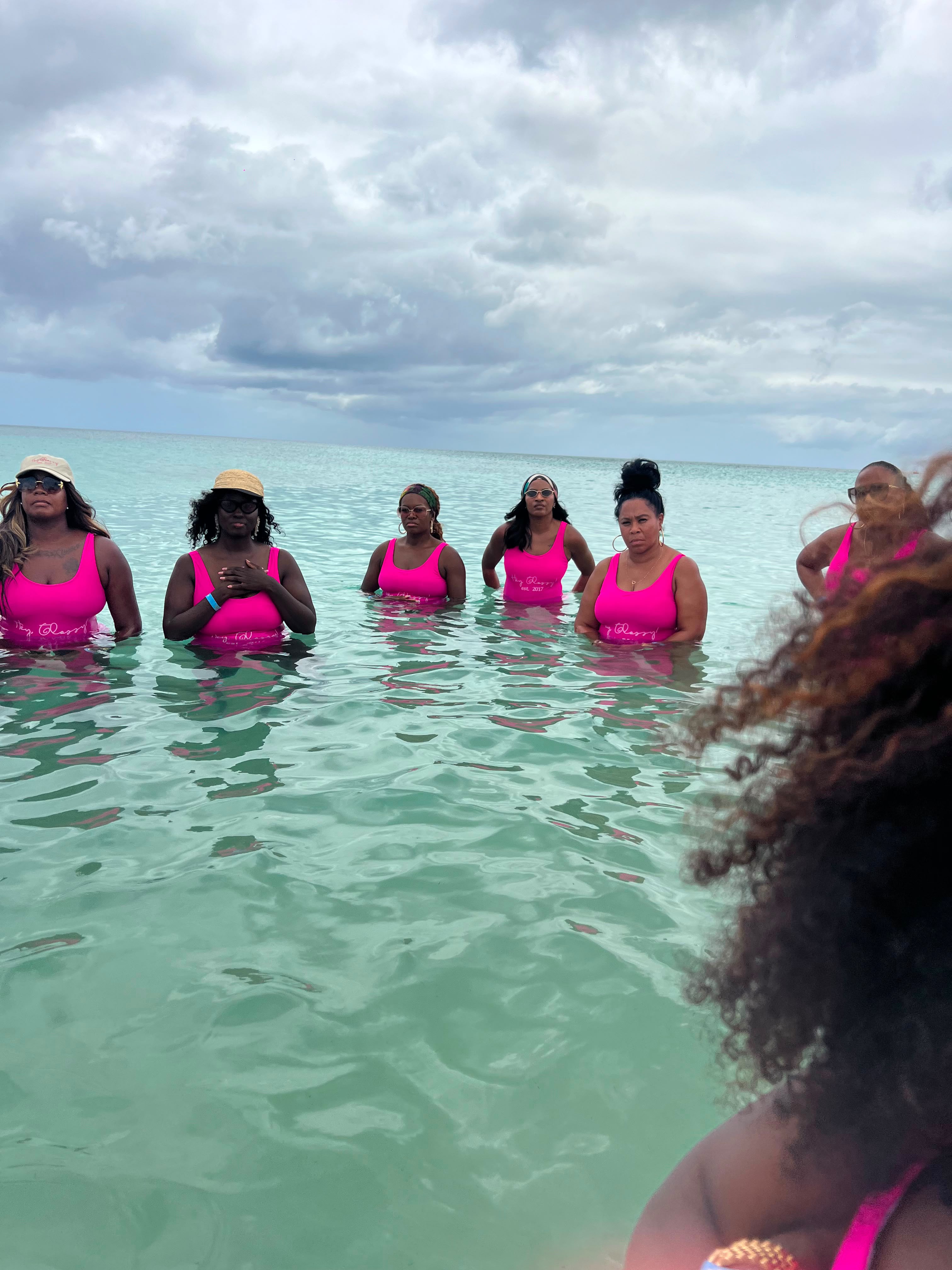 A group of women wearing pink swimsuits standing in a turquoise ocean under a cloudy sky.