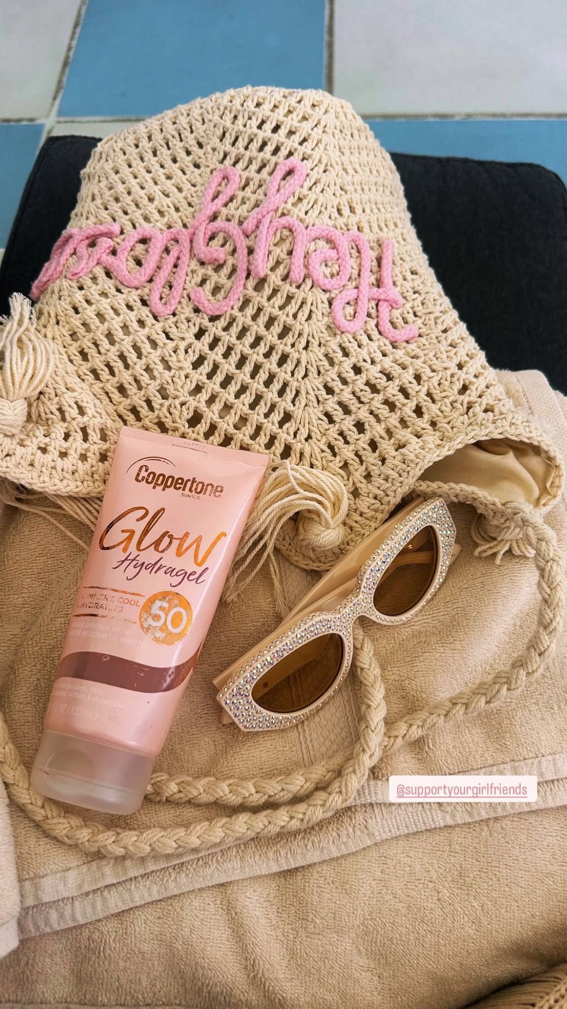 A crocheted sun hat with the word "Sunshine" embroidered on it, a Coppertone Glow hydragel sunscreen bottle, and a pair of rhinestone-encrusted sunglasses on a beach towel.