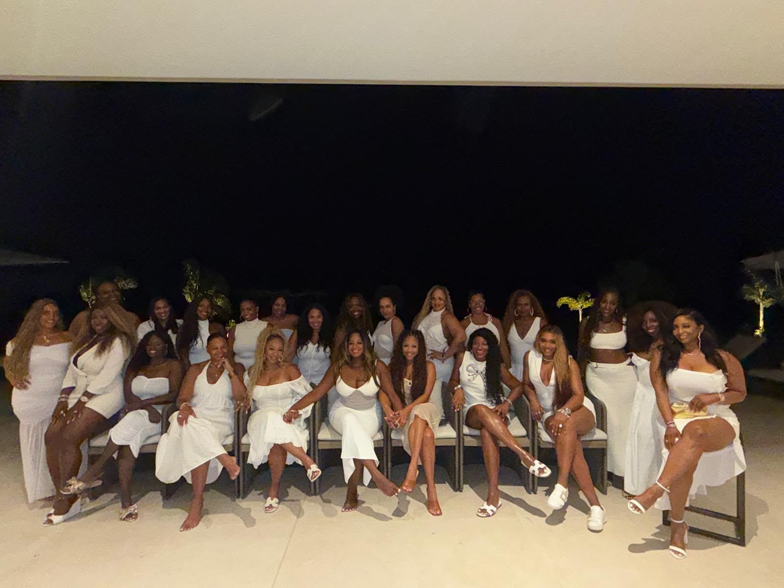 A large group of people, mostly women, wearing all-white outfits posing together in a dimly lit setting.