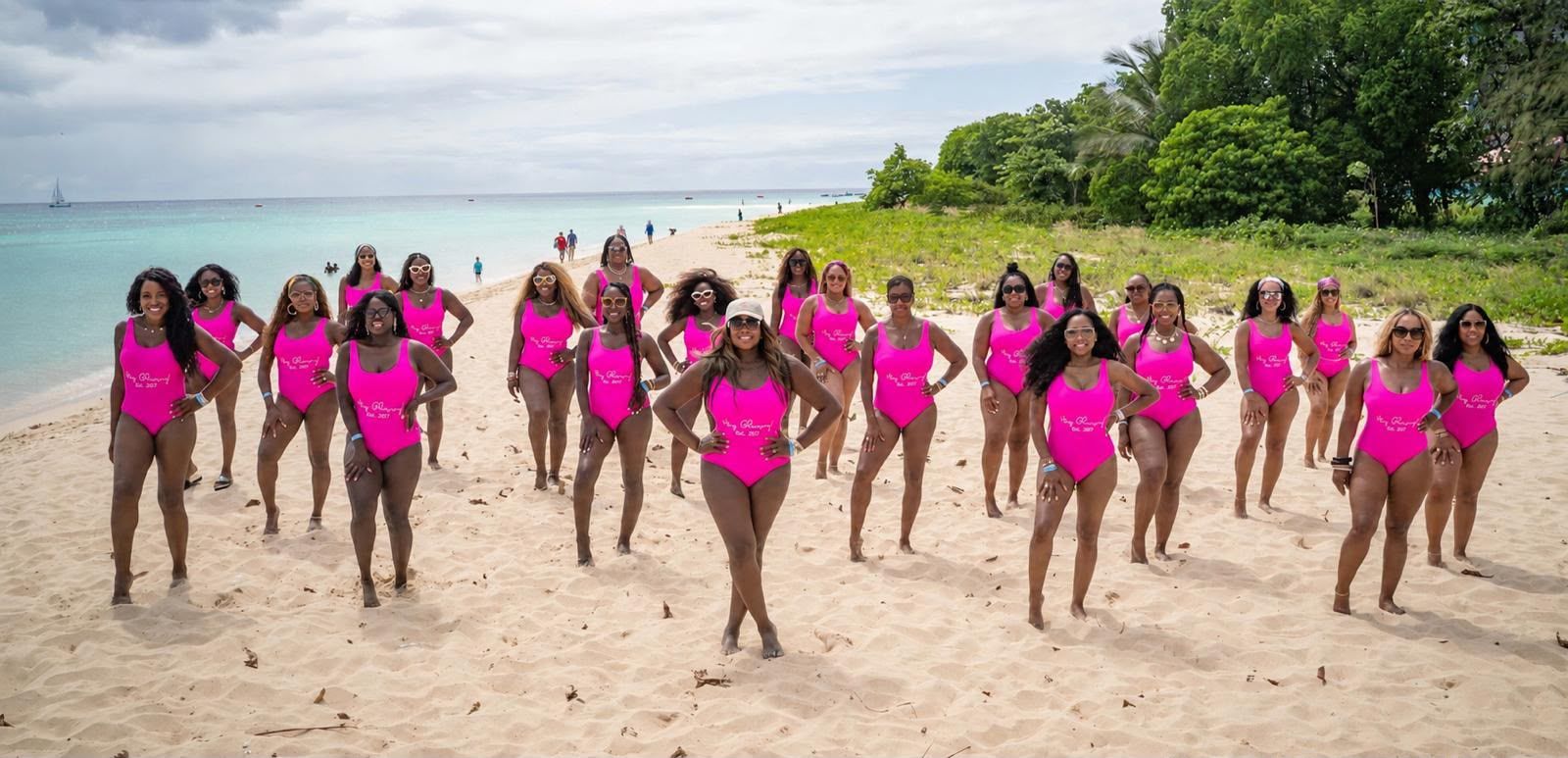 A group of women in bright pink swimsuits posing on a tropical beach with palm trees and clear blue water in the background.