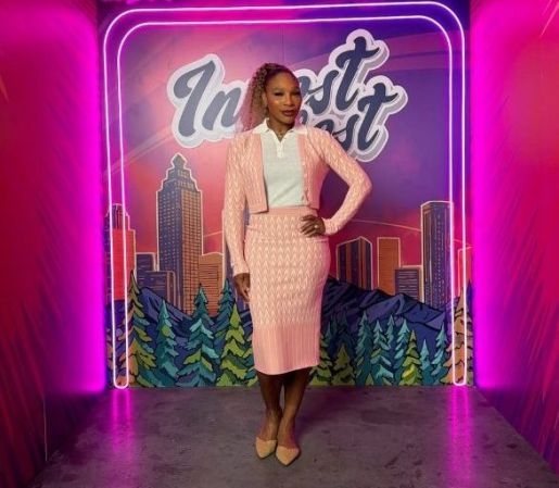 A woman wearing a pink outfit stands in front of a neon sign that says "In-est" against a backdrop of a cityscape and colorful clouds.