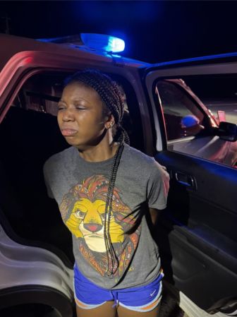 A person with braided hair sitting in a car at night, with blue and red police lights visible.
