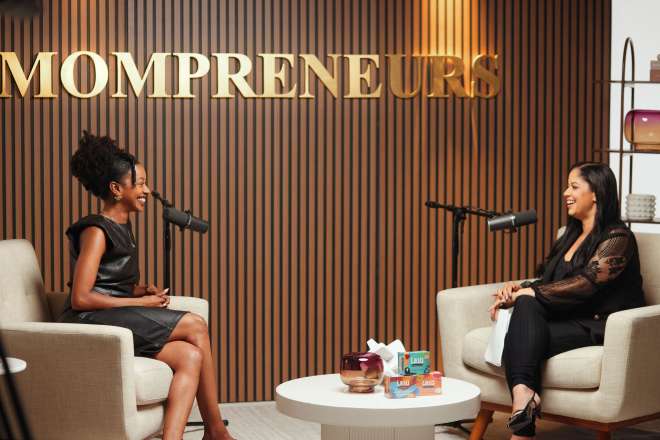 Two women sitting on chairs in a room with "MOMPRENEURS" sign on the wall behind them.