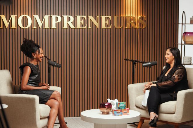 Two women sitting on chairs in a room with "MOMPRENEURS" sign on the wall behind them.
