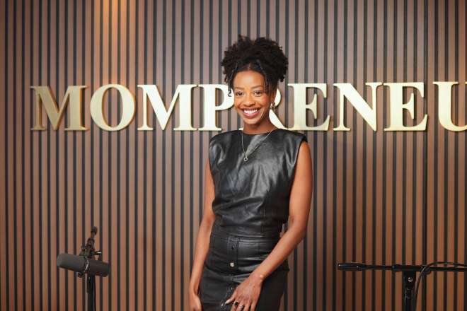 A smiling woman with curly hair standing in front of a sign that says "MOMPRENEU".